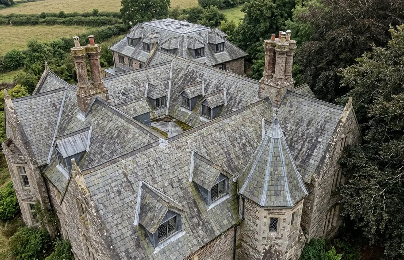 A large complicated house roof from the air with several pitches at different heights, three chimney stacks, dormers, valleys and mature trees pressing against one gable