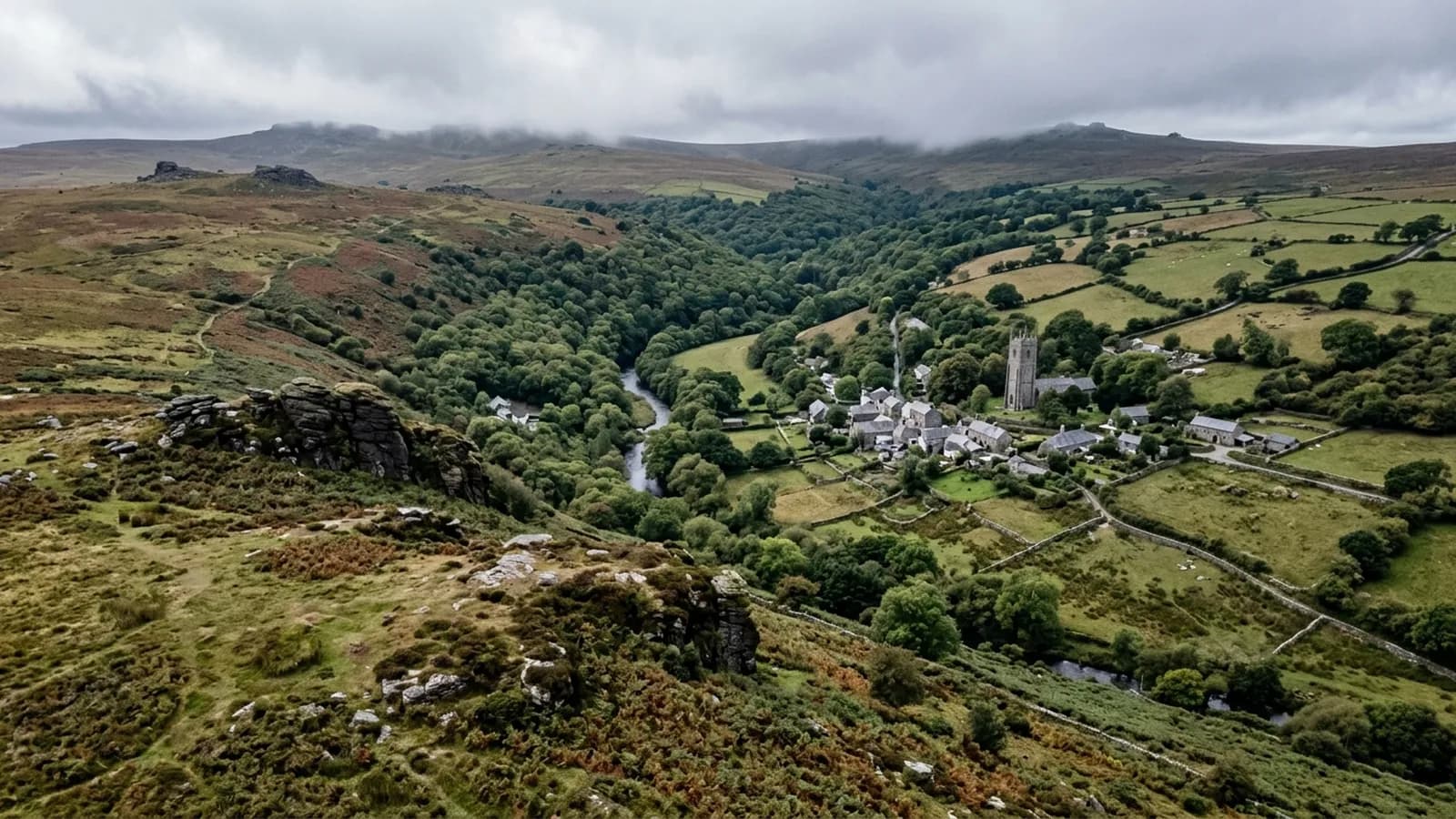 Open moorland with granite outcrops falling away to a wooded river valley with a stone village and a church tower, cloud sitting on the high ground