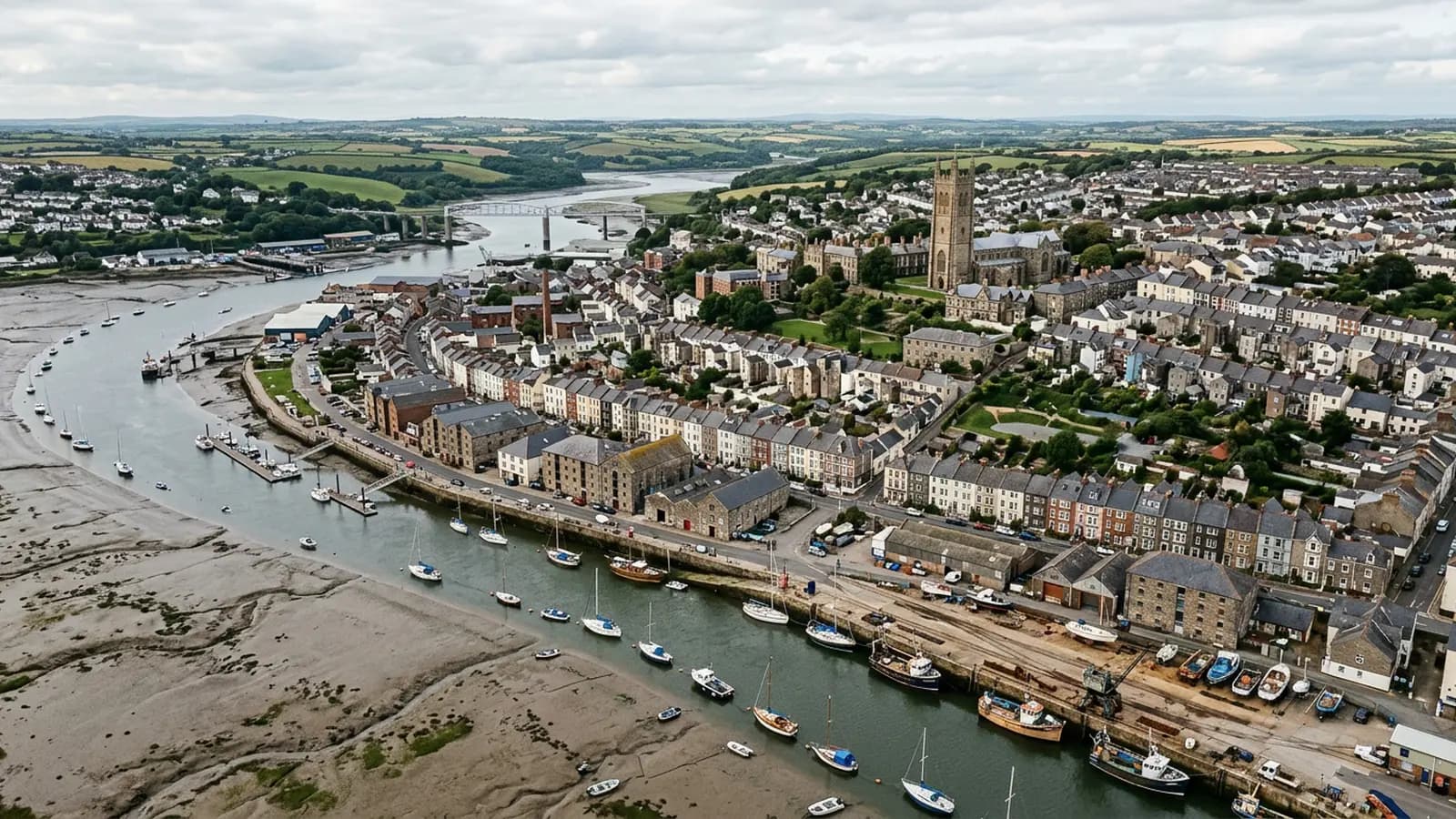 A tidal estuary city in Devon at half tide, mudflats and moored boats below a quayside of warehouses and terraces with a cathedral tower behind