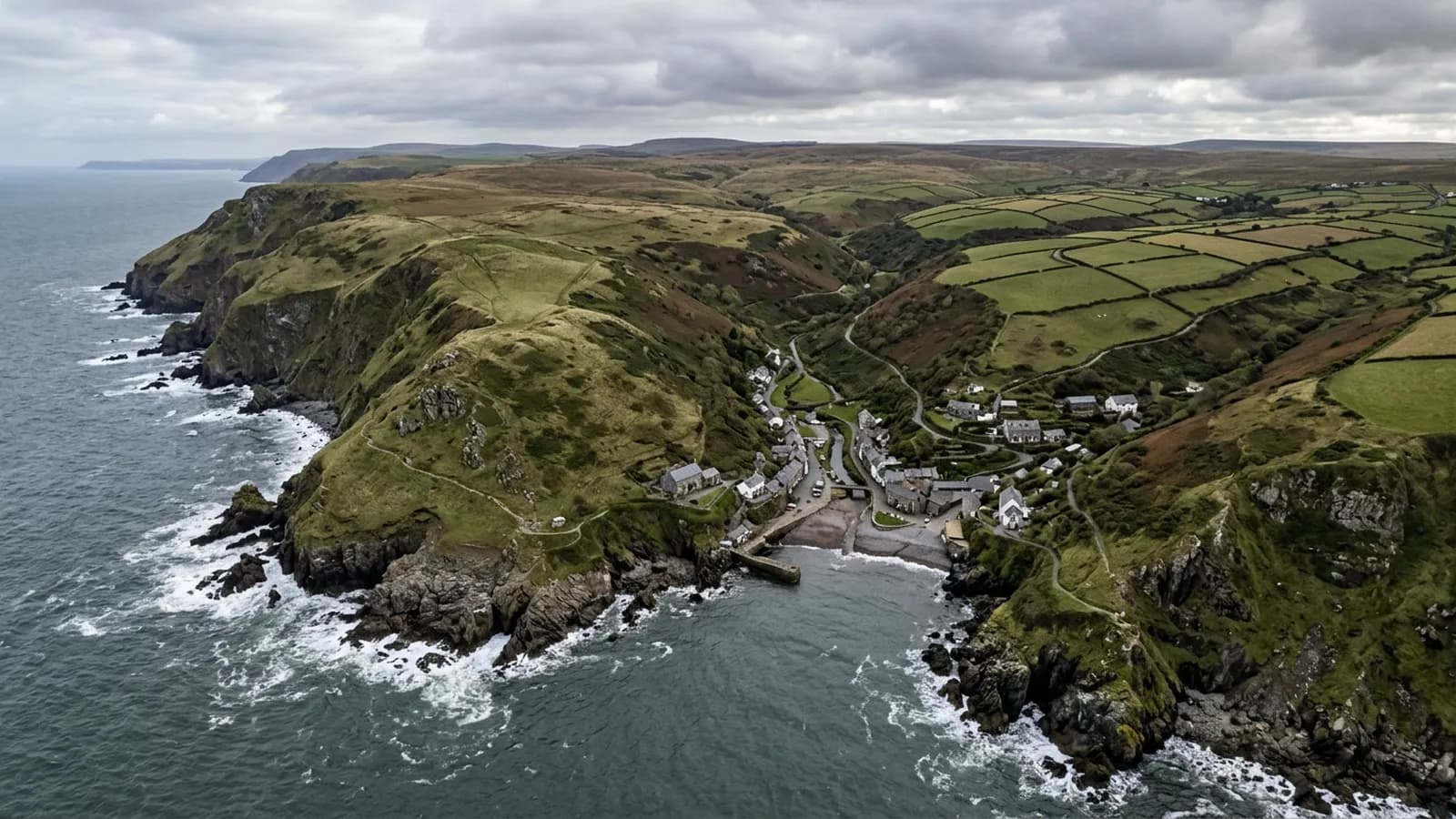 High grassy cliffs on the North Devon coast dropping to a rocky shore, with a combe running inland to a settlement of slate roofs